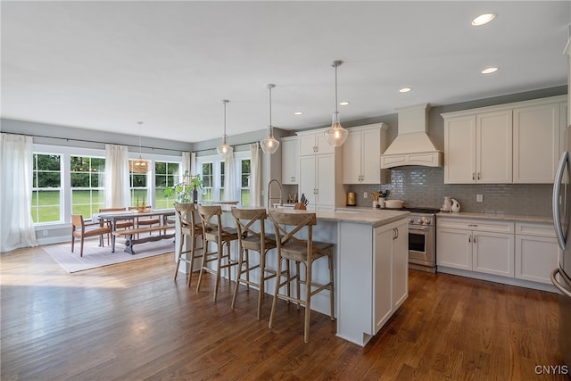 kitchen with pendant lighting, custom exhaust hood, a kitchen island with sink, dark wood-type flooring, and appliances with stainless steel finishes