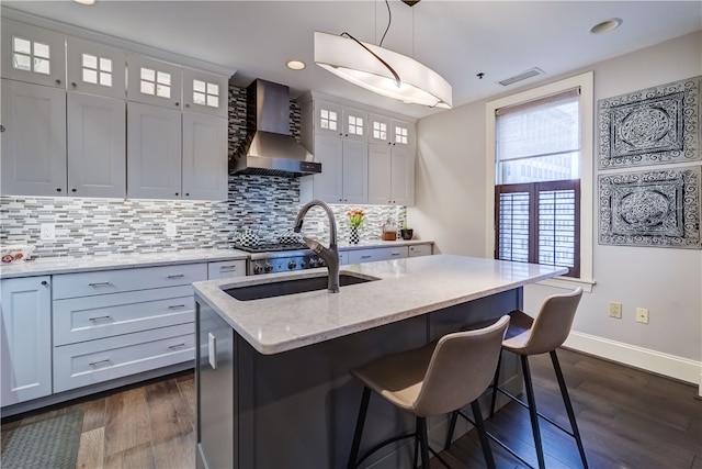 kitchen with light stone counters, a kitchen island with sink, white cabinets, hanging light fixtures, and wall chimney exhaust hood