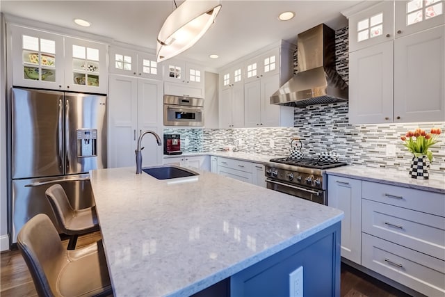 kitchen featuring an island with sink, white cabinets, wall chimney exhaust hood, stainless steel appliances, and decorative light fixtures