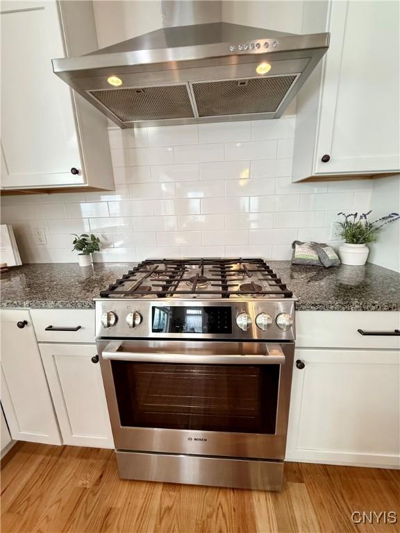 kitchen with stainless steel gas stove, wall chimney exhaust hood, dark stone counters, and white cabinets