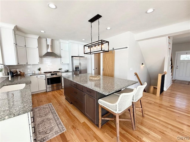 kitchen featuring wall chimney range hood, a barn door, appliances with stainless steel finishes, and white cabinets