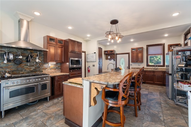 kitchen featuring a kitchen island with sink, wall chimney range hood, stainless steel appliances, light stone countertops, and decorative backsplash