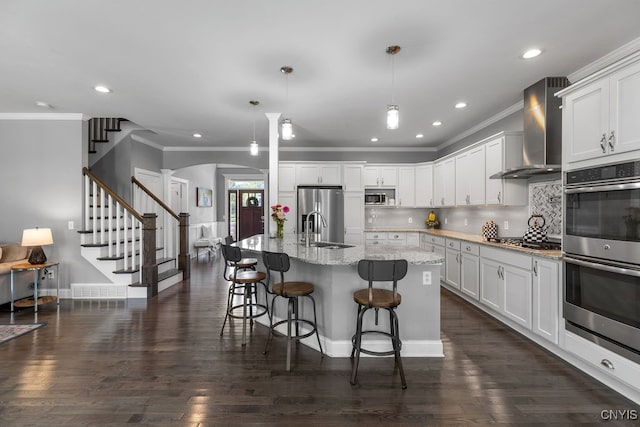 kitchen with white cabinets, wall chimney exhaust hood, stainless steel appliances, a center island with sink, and decorative light fixtures