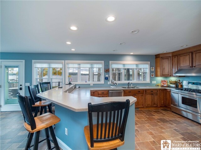 kitchen featuring a kitchen bar, under cabinet range hood, a sink, stainless steel appliances, and brown cabinetry