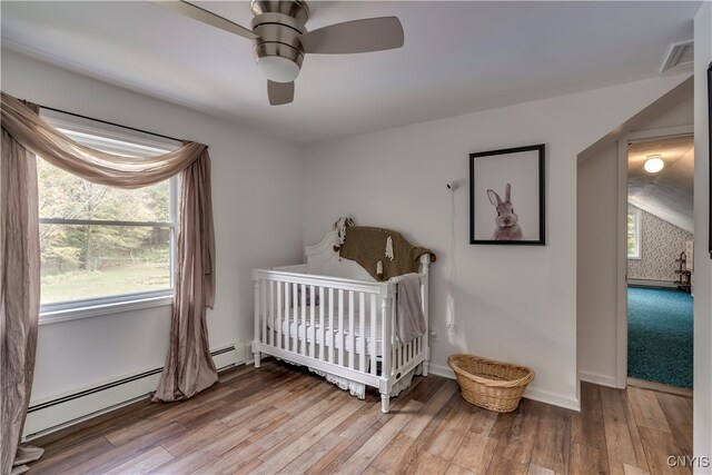 bedroom featuring ceiling fan, a nursery area, a baseboard heating unit, and wood-type flooring