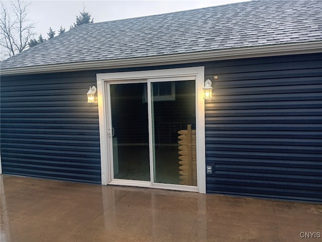 view of exterior entry with roof with shingles and a patio