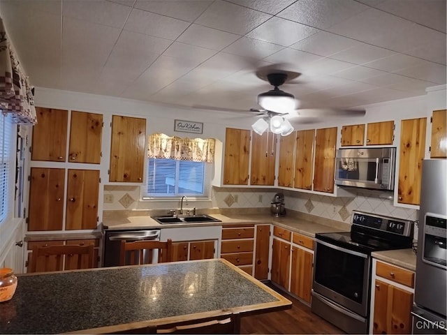 kitchen featuring stainless steel appliances, a sink, brown cabinetry, backsplash, and a ceiling fan