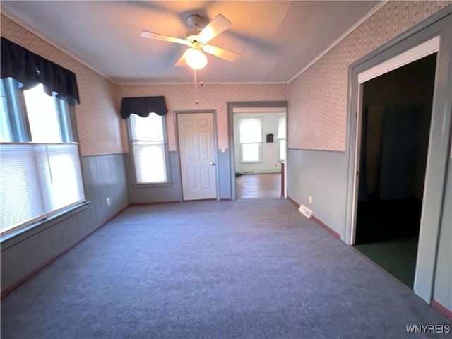 carpeted empty room with wallpapered walls, visible vents, a ceiling fan, a wainscoted wall, and ornamental molding