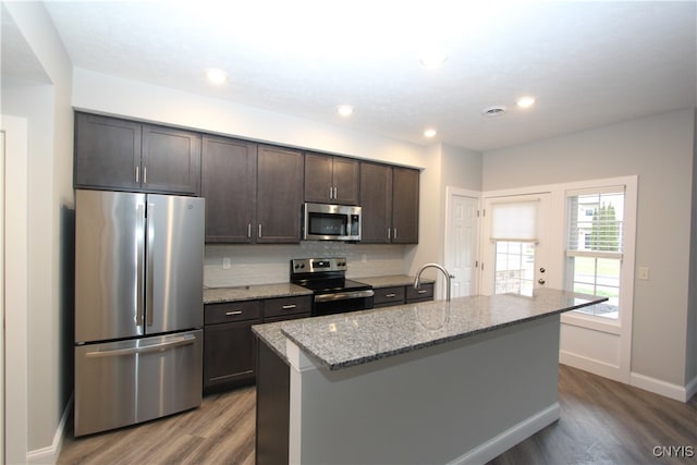 kitchen featuring an island with sink, appliances with stainless steel finishes, hardwood / wood-style flooring, light stone countertops, and dark brown cabinetry