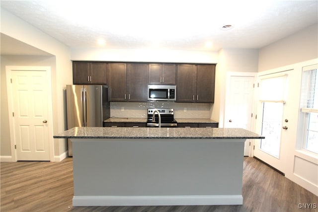 kitchen with appliances with stainless steel finishes, dark stone counters, dark hardwood / wood-style floors, and an island with sink