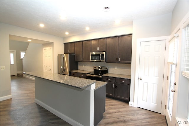 kitchen with appliances with stainless steel finishes, a kitchen island with sink, wood-type flooring, dark brown cabinetry, and light stone counters