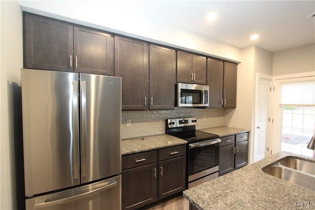 kitchen with tasteful backsplash, dark brown cabinets, light stone countertops, sink, and stainless steel appliances