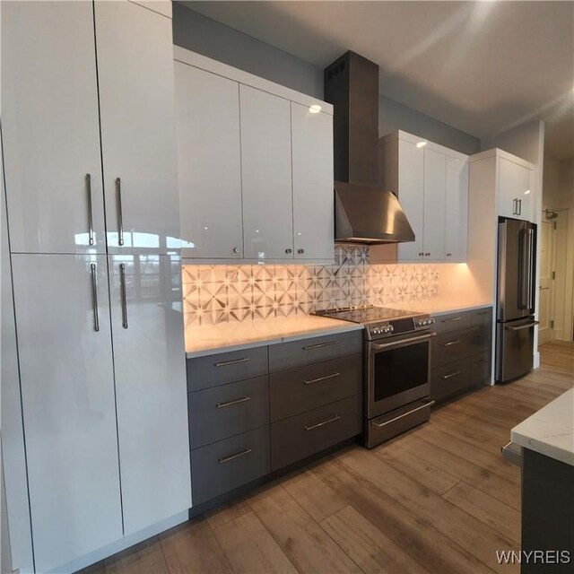 kitchen featuring wall chimney exhaust hood, white cabinetry, appliances with stainless steel finishes, hardwood / wood-style flooring, and backsplash