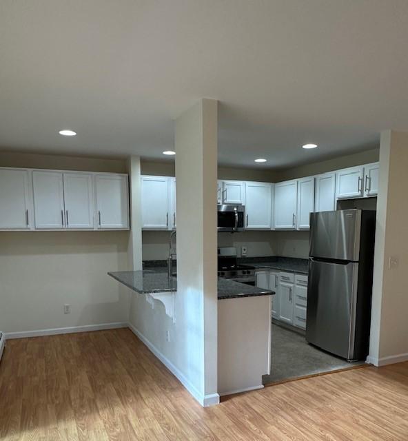 kitchen with kitchen peninsula, appliances with stainless steel finishes, light wood-type flooring, dark stone countertops, and white cabinetry