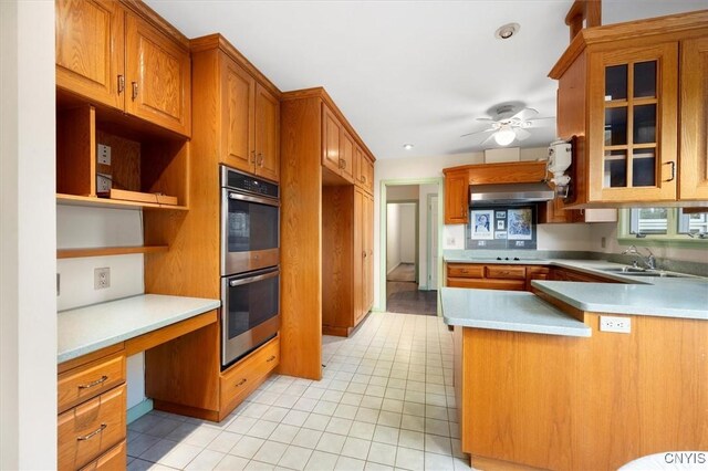 kitchen with sink, ceiling fan, double oven, built in desk, and wall chimney exhaust hood