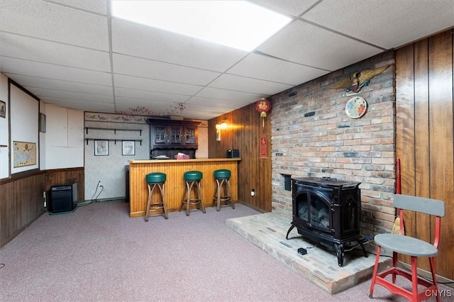 bar featuring a wood stove, light carpet, a drop ceiling, and wood walls