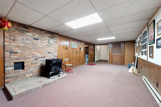 basement with wood walls, a drop ceiling, a wood stove, carpet, and a baseboard heating unit