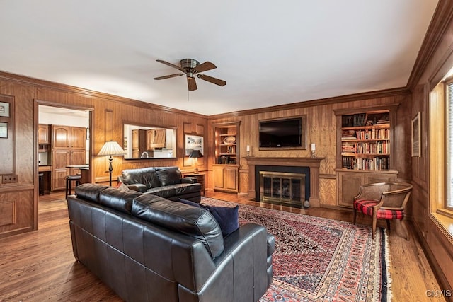 living room with wooden walls, wood-type flooring, ornamental molding, built in shelves, and ceiling fan