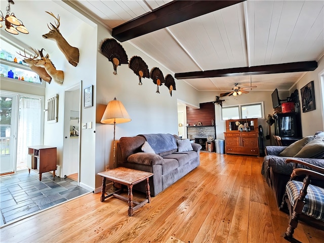 living room featuring a stone fireplace, beam ceiling, wood-type flooring, and ceiling fan