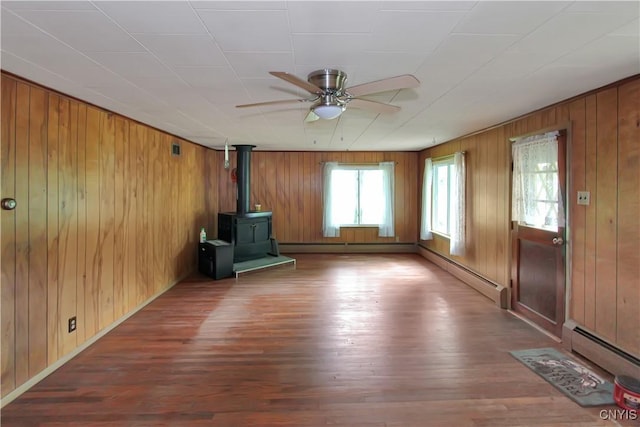 6736 Pine Grove Rd, Watson, NY 13343 - unfurnished living room with a wood stove, ceiling fan, wood finished floors, wooden walls, and a baseboard radiator
