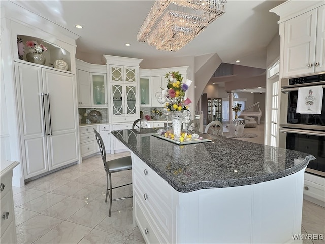 kitchen with stainless steel double oven, a kitchen island with sink, dark stone countertops, a notable chandelier, and white cabinetry