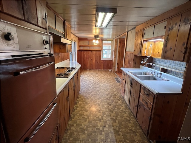 kitchen featuring stainless steel gas cooktop, oven, wooden walls, sink, and ceiling fan