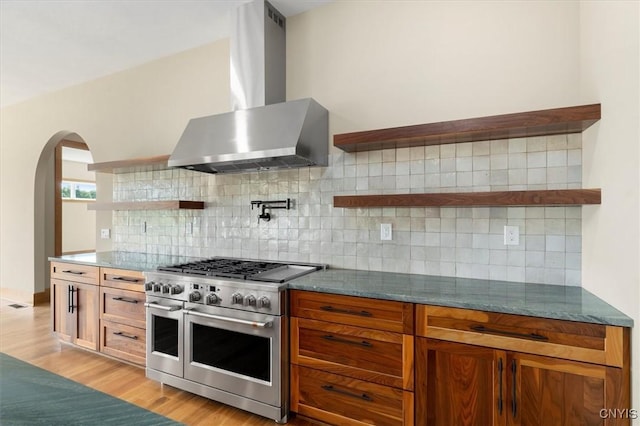 kitchen featuring range with two ovens, wall chimney exhaust hood, light hardwood / wood-style floors, backsplash, and dark stone countertops