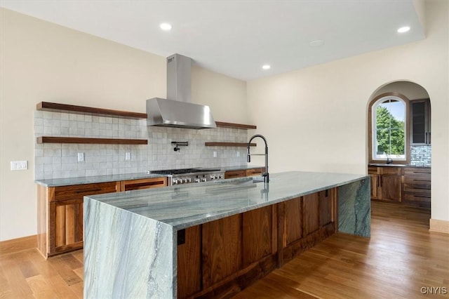 kitchen with light stone countertops, wall chimney range hood, decorative backsplash, light wood-type flooring, and sink