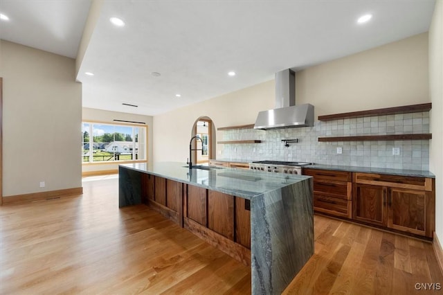 kitchen with a center island with sink, sink, light hardwood / wood-style flooring, wall chimney range hood, and backsplash