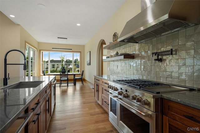 kitchen with wall chimney exhaust hood, double oven range, light hardwood / wood-style floors, dark stone countertops, and sink