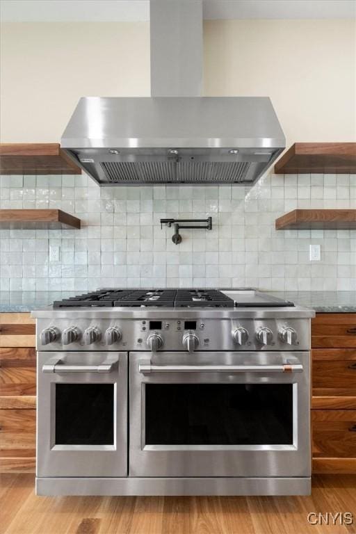 kitchen with light wood-type flooring, double oven range, light stone counters, decorative backsplash, and wall chimney exhaust hood