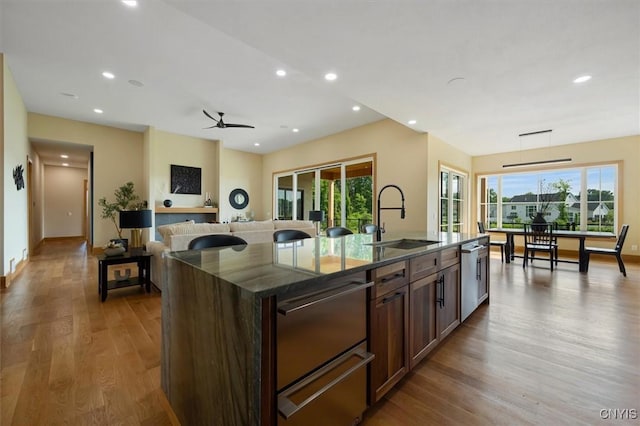 kitchen featuring hardwood / wood-style flooring, a kitchen island with sink, dark stone counters, ceiling fan, and sink