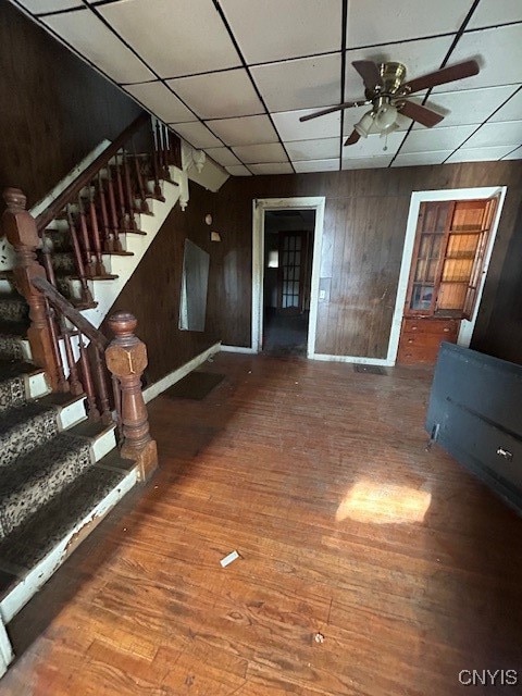 interior space featuring a drop ceiling, wooden walls, wood-type flooring, and ceiling fan