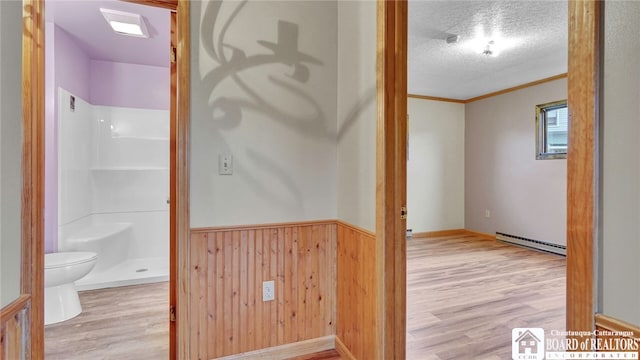 hallway with a textured ceiling, a baseboard heating unit, crown molding, wooden walls, and light wood-type flooring