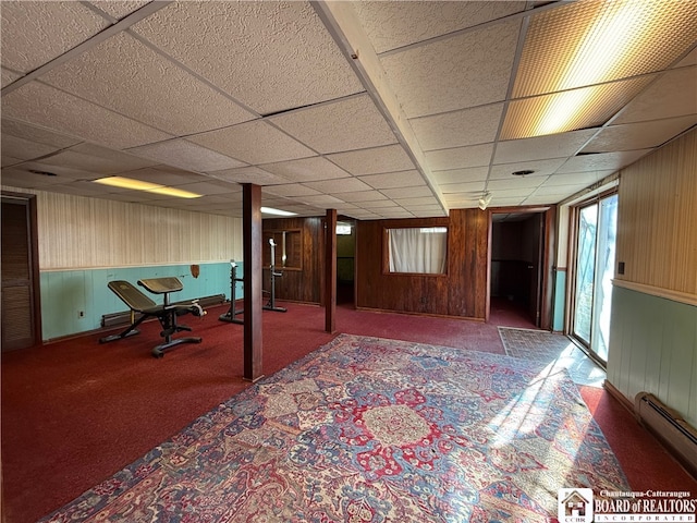 interior space with a drop ceiling, a baseboard radiator, carpet floors, and wood walls