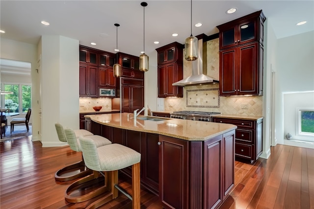 kitchen with tasteful backsplash, wall chimney exhaust hood, decorative light fixtures, a kitchen island with sink, and dark wood-type flooring