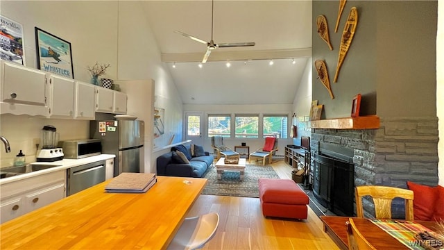 living area with ceiling fan, high vaulted ceiling, a stone fireplace, light wood-style flooring, and track lighting