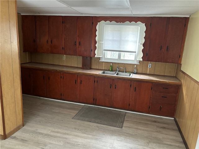 kitchen featuring light wood-type flooring, a drop ceiling, wooden walls, sink, and tile counters