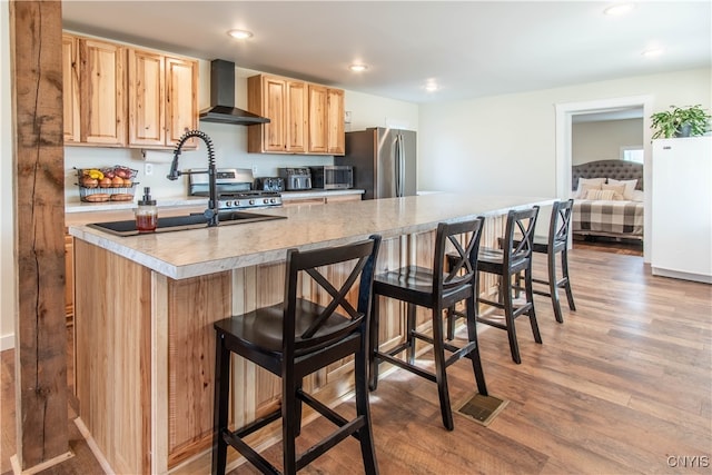 kitchen with a breakfast bar, wall chimney range hood, light wood-type flooring, light brown cabinetry, and appliances with stainless steel finishes