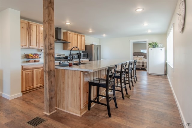 kitchen featuring stainless steel appliances, wall chimney range hood, light hardwood / wood-style floors, light brown cabinetry, and a center island with sink