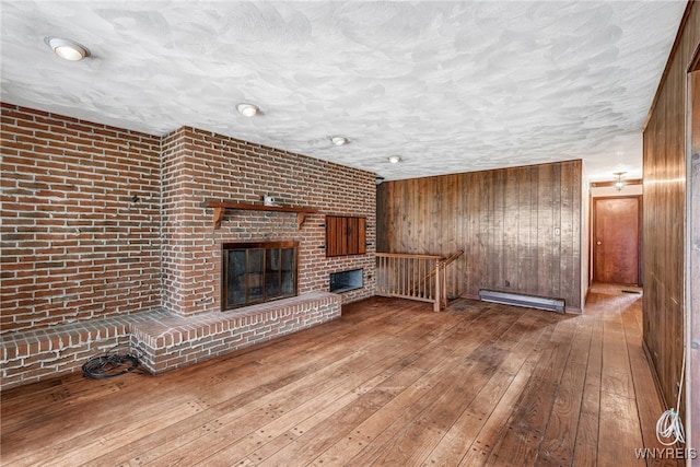 unfurnished living room featuring a brick fireplace, a textured ceiling, wooden walls, wood-type flooring, and a baseboard radiator