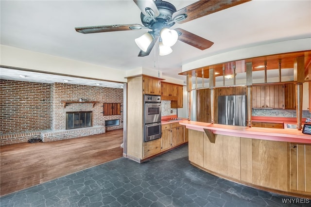 kitchen with ceiling fan, dark wood-type flooring, a brick fireplace, brick wall, and appliances with stainless steel finishes