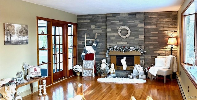 living area with a stone fireplace, a wealth of natural light, wood-type flooring, and a baseboard radiator