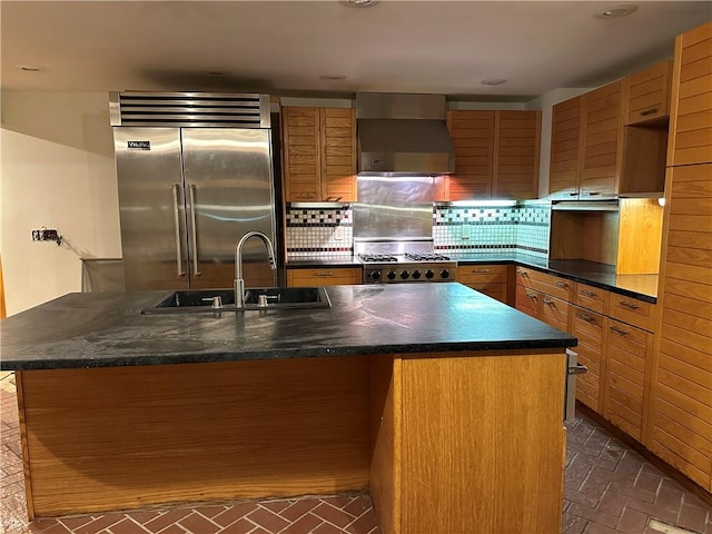 kitchen featuring backsplash, stainless steel appliances, sink, wall chimney range hood, and a center island with sink