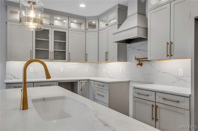 kitchen featuring custom range hood, decorative light fixtures, gray cabinetry, light stone counters, and decorative backsplash