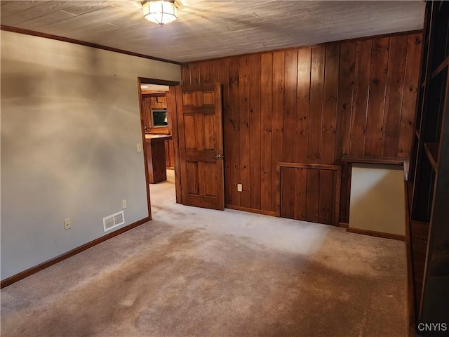 empty room with crown molding, light colored carpet, visible vents, wood walls, and baseboards