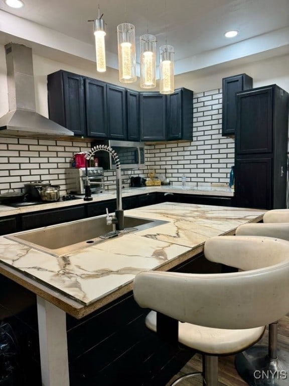 kitchen with hanging light fixtures, wall chimney range hood, tasteful backsplash, a kitchen bar, and a kitchen island with sink