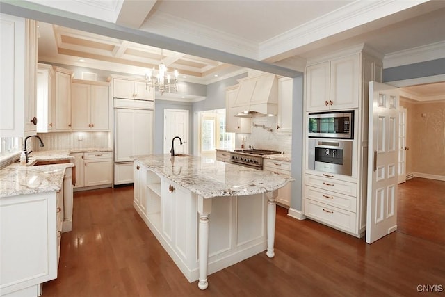 kitchen with custom exhaust hood, coffered ceiling, light stone countertops, a center island with sink, and built in appliances