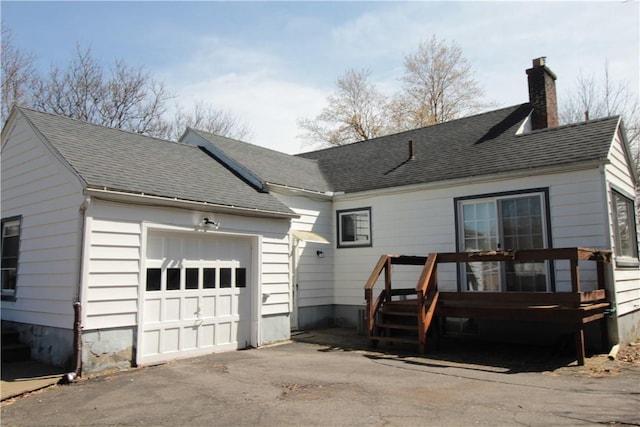 back of house featuring an attached garage, a chimney, a wooden deck, driveway, and roof with shingles