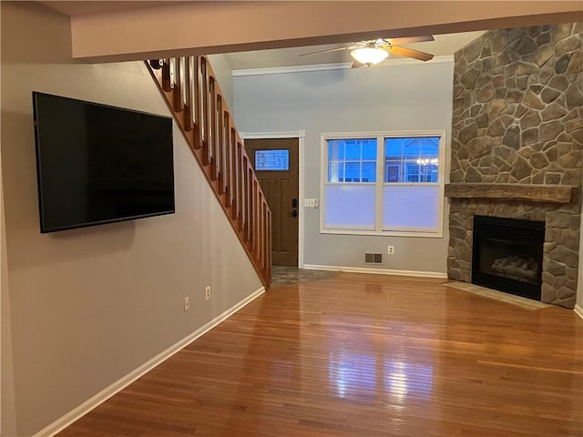 unfurnished living room featuring ceiling fan, a stone fireplace, wood-type flooring, and crown molding